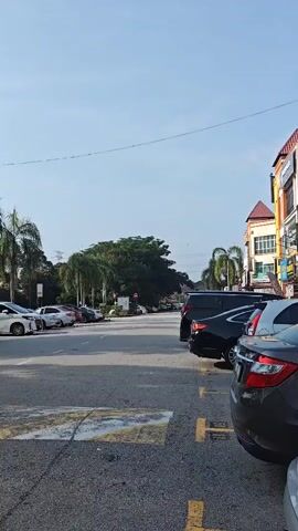 A parking lot is depicted with a clear blue sky above. Several cars are parked on the left side of the road, and more cars are parked on the right side. Trees and buildings are visible in the background. The asphalt of the parking lot is marked with yellow lines.