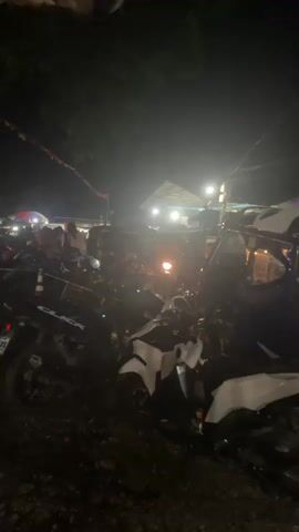 The video shows a crowded scene at night, likely an outdoor market or event based on the lighting and the presence of multiple vendor stalls. The foreground is filled with parked motorcycles, mostly dark in color. A blue and white vehicle, possibly a tuk-tuk or similar public transport, is visible on the right side. In the background, there's a gathering of people, illuminated by bright overhead lights. A small building with a thatched roof can be seen further in the background. There is a sense of activity and bustling movement.