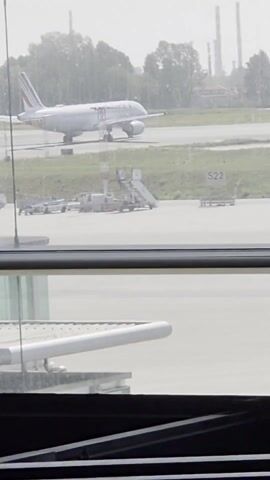 The video presents a daytime view of an Air France airplane stationary on an airport runway, observed through a window. The plane, white with the Air France logo and red and blue accents on the tail, is positioned to the left of the frame. Adjacent to the plane are airport ground support equipment, including a set of stairs. A sign reading "S22" is visible in the background. In the distance, green trees and industrial chimneys are discernible, contributing to the overall environment. The focus is sharp on the scene outside the window, while the immediate foreground contains parts of the window frame, slightly obscuring the view. The scene is uniformly lit, providing clear visibility.