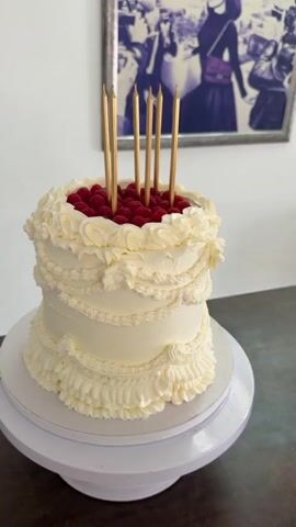 A white cake adorned with cream frosting and fresh raspberries sits on a white cake stand. The cake is decorated with ruffles and piping along its sides. Five gold candles stand upright in the center of the cake, nestled among the raspberries. The cake is positioned in front of a wall with a framed picture hanging above it. The picture appears to be a vintage black and white photograph featuring several individuals wearing elaborate clothing. The scene is lit by natural light, casting soft shadows. The camera slowly rotates around the cake, offering a 360-degree view.