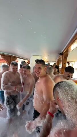 A group of people, mostly men, are enjoying a foam party inside what appears to be a boat. The boat's interior has a light-colored ceiling and wooden beams, with windows along the side revealing bright daylight and water, suggesting it's moving on the sea. Foam covers the floor and many of the attendees, who are largely shirtless or in swim attire. Some individuals have significant amounts of foam on their hair and bodies, indicating an active foam machine. People are seen dancing, mingling, and moving through the thick layer of foam. The atmosphere appears lively and celebratory. The event is taking place on a sunny day, likely during a boat trip in the Menderes region of Turkey, as per the provided context.