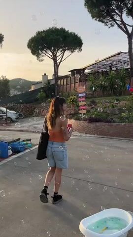 A woman with brown hair, wearing an orange crop top, denim shorts, and black sneakers, stands outside. She holds a red and white striped popcorn container in her left hand. Bubbles float around her. In the background, there is a building, trees, and a hill. The woman turns to her right, smiles, and waves her right arm. A black tote bag hangs from her left shoulder. She appears to be standing on a paved surface.