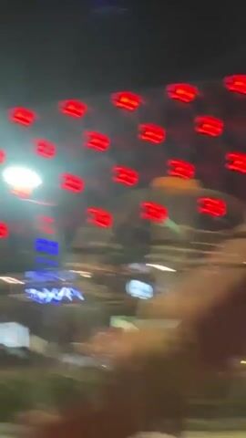 The video, taken from a moving car at night, shows the exterior of a brightly lit shopping center called "RÜYA OUTLET". Palm trees are visible in front of the building, illuminated by the shopping center's lights. The building itself features a modern design with multiple arches and a wide storefront. Cars and other vehicles can be seen in the parking area. The perspective is from the passenger side window of the moving car.