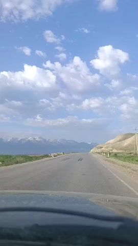 The video shows a first-person view from inside a vehicle, traveling on a paved road. The road stretches ahead, leading towards a mountain range in the distance. The sky is bright blue with scattered white clouds. On the left side of the road, there is a field, and further in the distance, snow-capped mountains are visible. On the right side of the road, there is a gently sloping hill covered in dry grass and some sparse vegetation. White roadside posts are visible along the right edge of the road. The road appears to have tire marks from previous vehicles. The lighting suggests it is daytime, possibly late afternoon due to the angle of the sun.