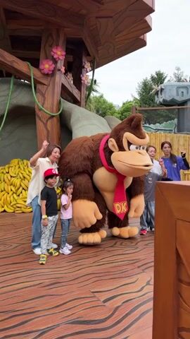 This image depicts a group of seven people—two adult women, an adult man, and four children—posing for a photograph with a large Donkey Kong mascot character. The setting is an outdoor stage area with a wooden floor, designed to resemble a jungle or treehouse, complete with a prominent wooden structure and a large pile of yellow bananas on the left.

The Donkey Kong character, with its brown fur and large features, stands centrally, wearing a red tie emblazoned with "DK" in yellow letters. The people around it are smiling and engaging with the character; one adult woman flexes her arm, while another adult woman and a child make peace signs.

In the background, green foliage and bamboo fencing are visible, along with a distinctive stone castle, which appears to be Hogwarts Castle from The Wizarding World of Harry Potter. This suggests the location is Universal Studios Japan in Osaka. The scene is brightly lit, indicating fair weather during the daytime. The overall mood conveyed is one of joy and excitement, typical of a theme park visit.