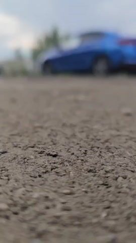 The video begins with a close-up, low-angle shot of a textured, grayish-brown surface, possibly asphalt or dirt. The foreground is in sharp focus, revealing small pebbles and a rough texture. In the background, slightly out of focus, is a blue car. The car appears to be parked, and its rear is visible. The sky above is overcast with diffused light. As the video progresses, the camera remains stationary, and the background elements show a slight blur, indicating a very shallow depth of field. There are no people visible in the frame. The environment appears to be an outdoor setting, possibly a parking lot or a roadside area.