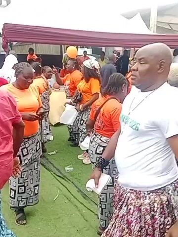 The video shows an outdoor gathering. Several people are present, most of whom are wearing orange shirts and patterned skirts or wraps. Some individuals are holding balloons, and a few are holding white flags on sticks. The ground is covered with green turf. In the background, there are tents and a building. Some people appear to be engaged in some sort of organized activity or performance.