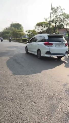 The video shows a street scene with traffic. A white SUV is driving on the road, followed by a motorcycle. The motorcycle has a rider wearing a dark jacket and helmet. The road appears to be asphalt and is somewhat worn. There are trees and buildings visible on the side of the road. The sky is overcast. The video appears to be shot from another vehicle, possibly a motorcycle, as indicated by the handlebars visible in the foreground. The camera is in motion, following the white SUV and the motorcycle.