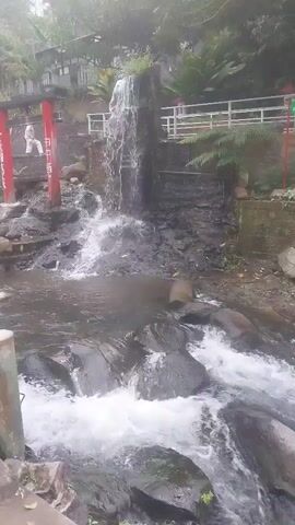 The video captures a flowing waterfall cascading over rocks into a stream. There are red torii gates on the left side of the frame, and a white railing is visible along the top right. A person in white is standing on a platform near the top of the waterfall. The environment appears to be natural, with rocks, trees, and vegetation. The water is churning and creating white foam as it flows over the rocks.