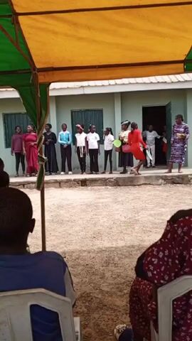 From a partially shaded perspective, the video shows a group of individuals, seemingly students and teachers, assembled outside a light green building. A yellow and green canopy offers a partial view of the outdoor gathering. The students, dressed in a variety of casual and formal attire, stand in a line on a slightly elevated platform in front of the building. A teacher or speaker stands to the right, holding a microphone and addressing the group. Other individuals are present both on the platform and near the open doorway of the building. In the foreground, the backs of several seated attendees are visible. The ground is covered with gravel or small stones. The overall setting suggests an outdoor event, possibly a school function or gathering.