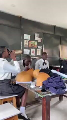 The scene captures a classroom setting where three students are engaged in an art project involving large, light brown gourds. The girl to the left is seated on a wooden stool, and is holding a metallic water bottle. She is dressed in a white collared shirt and a dark skirt.  Next to her, a male student is bent over one of the gourds, seemingly applying paint or some other substance with a small brush. To the right, another student is also seated at the table. The table they are working at is wooden, stained dark. On the table are the gourds, as well as some art supplies and cloth material. The background features a bulletin board covered with various pinned items. The classroom has natural lighting, and the floor appears to be tiled.