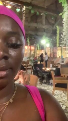 A close-up video of a woman with dark skin wearing a bright pink headband and a pink top. She is looking at the camera and pursing her lips. She has long eyelashes and is wearing two gold necklaces. The background is out of focus and appears to be an indoor dining area with tables and chairs. Some people are visible in the background, including a man and a woman standing near a table with a fire pit. There are hanging plants and lights in the ceiling, creating a decorative atmosphere. The flooring is a mosaic of irregular tiles. The lighting is dim, suggesting it is evening or nighttime.