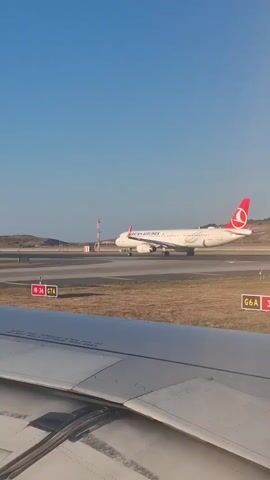 This is a POV shot from inside an airplane, looking out the window at another airplane on the tarmac. The airplane in the foreground has a portion of its wing visible, with flaps and leading-edge devices. The airplane in the distance is a Turkish Airlines plane, white with a red tail fin featuring the Turkish Airlines logo. It is on a runway or taxiway, with other markings and structures visible in the background, including runway lights and signage. The sky is clear and blue. The overall scene depicts an airport environment with aircraft on the ground.