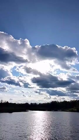 This is a vertical video clip of a natural scene. The primary subject is the sky, which is filled with a dynamic display of clouds. The sun is partially obscured by these clouds, creating dramatic shafts of light that break through. The light reflects brightly on the surface of a body of water that occupies the lower portion of the frame.

In the distance, a treeline of dark green, silhouetted trees forms the horizon. The overall impression is one of a vast, open landscape.

The time of day appears to be either late afternoon or early evening, given the angle of the sun and the quality of the light. The weather seems to be partly cloudy, with both bright sunshine and darker, more ominous-looking clouds present.

There is no discernible human presence or man-made objects apart from what might be a very distant, thin structure on the far right horizon, barely visible against the sky.

The mood of the scene is peaceful and somewhat dramatic, evoked by the interplay of light and shadow in the sky and on the water. The scene is likely a lake or a wide river in a natural, possibly remote, location. The provided location context of "1-Go Maya, Russia" suggests a rural or wilderness setting.

No visible text is present in the media file.