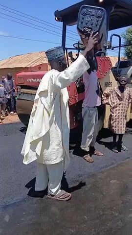 A group of people, including men, women, and children, are gathered on a paved road. A large yellow road roller, a Dynapac, is present. Several individuals are holding posters or signs. One man in a white robe and cap is holding up a sign and appears to be gesturing. Another man is wearing a pink shirt and grey pants. There are also individuals in security uniforms and a person in a high-visibility vest. The environment appears to be outdoors on a sunny day, with buildings and trees visible in the background. The road surface is freshly paved asphalt.