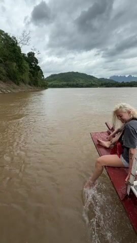 A woman with blonde hair sits on the edge of a boat, trailing her feet in the murky brown water. She is wearing a dark t-shirt and red shorts. Her legs are bare and her feet are splashing in the river. The boat appears to be moving, as evidenced by the water splashing around her feet. The riverbank on the left is lined with green trees and foliage. In the background, a hazy mountain range is visible under a cloudy sky. The overall atmosphere is one of a relaxed boat trip on a river.