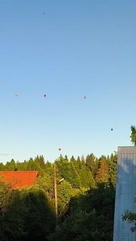 The video shows a clear blue sky with several hot air balloons in the distance. Below the balloons, a treeline of green trees is visible, with the roof of a building and a utility pole in the foreground. The hot air balloons are colorful and appear to be spread out across the sky.