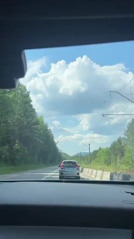 This is a first-person point-of-view video taken from inside a car driving on a paved road through a wooded, mountainous area. The time of day appears to be midday with bright sunshine and scattered cumulus clouds in a blue sky. The road has white lane markings and a concrete barrier on the right side. There are tall pine trees lining the left side of the road, and more trees and hills in the distance. Overhead power lines for what appears to be a railway are visible on the right.

The car in front is a light-colored Honda hatchback. Further ahead, a white truck with a crane arm is visible, and in the distance, another dark-colored car is approaching.

The overall mood is one of travel and exploration through a scenic natural landscape. The visible text on the license plate of the Honda is too blurry to be deciphered. The location context provided is Ust-Mana, Russia.