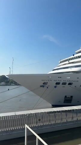 A large white cruise ship named "SEVEN SEAS MARINER" is docked next to a gangway. The ship is tied to the dock with ropes. The sky is clear and blue. A person is visible on the gangway.