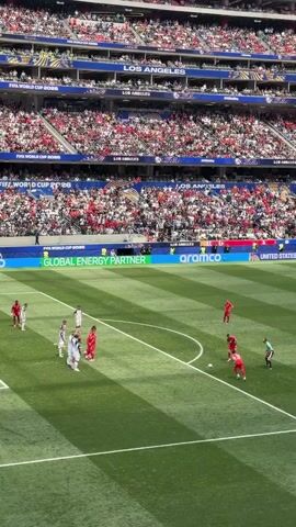 A soccer game is in progress. The field is green with white lines. A goalie in a yellow jersey is in the penalty box. Several players in red and white jerseys are on the field. The stands are full of spectators wearing red and white. A banner at the bottom of the stands reads "ARAMCO GLOBAL ENERGY PARTNER." The sky is overcast.
