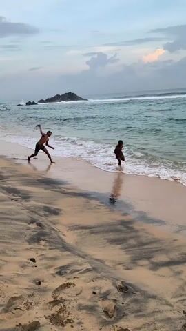 On a beach with streaks of black sand, two dark-skinned individuals are running along the shoreline. One person is visible closer to the camera, sprinting with their arms outstretched towards the ocean. Their reflection is visible on the wet sand. The ocean has turquoise waves, and there is a rocky island in the distance under a cloudy sky.