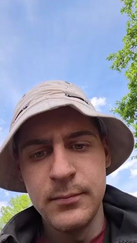 A man with a bucket hat stands outdoors under a blue sky with scattered clouds. He is speaking to the camera, making hand gestures. A tree with green leaves is visible in the upper right of the frame.