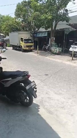 A person riding a red three-wheeled motorcycle with a red cargo bed is captured in motion from behind and to the side. The motorcycle is moving from the left to the right of the frame. The person is wearing a dark shirt, dark pants, and a black helmet, and is looking forward. The motorcycle has a visible license plate. In the background, a yellow truck with a white cargo box is parked, also facing right. Several other motorcycles are parked on the side of the road. The scene appears to be a street with shops and trees lining the sides. The weather is sunny with shadows visible on the road.