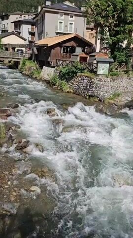 This video showcases a picturesque view of Ponte di Legno, Italy, on a bright, sunny day. The scene opens with a close-up of a rapidly flowing river, its clear water churning into white rapids as it cascades over rocks. The riverbanks are lined with natural stone and lush green vegetation.

As the camera slowly pans upwards and to the right, it reveals a charming Alpine town nestled along the river. Traditional multi-story buildings with pitched roofs, balconies, and warm-colored facades (such as orange, yellow, and beige) are prominent, displaying typical European mountain architecture. A small wooden cabin-like structure is visible near the river on the left bank.

A stone bridge spans the river, and a dark-colored SUV, possibly a Mercedes G-Wagen, is parked on it. A paved road runs through the town, and in the distance, two small figures of people can be seen walking, indicating a quiet and peaceful atmosphere.

The most striking element of the scene is the backdrop of majestic mountains. Their rugged peaks, partially covered in snow, dominate the horizon under a clear blue sky dotted with fluffy white clouds. The contrast between the vibrant green of the lower slopes and the pristine white snow on the higher elevations is stunning. The time of day appears to be midday or early afternoon, with abundant sunlight illuminating the entire landscape. No legible text is visible on any signs or objects in the video.