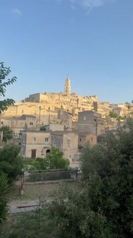 A vertical video shows an ancient European city with buildings constructed of tan stone. The architecture is densely packed, with structures built on a hillside and appearing to cascade down. Many of the buildings have arched windows and doorways. A tall bell tower, also made of tan stone, rises above the cityscape. Green foliage is visible in the foreground, framing the view of the city. The sky is a clear, pale blue. The lighting suggests it is either early morning or late afternoon, with the sun casting warm tones on the buildings. The video is a zoom-in, starting with a wider view and gradually focusing on the bell tower and the surrounding architecture.