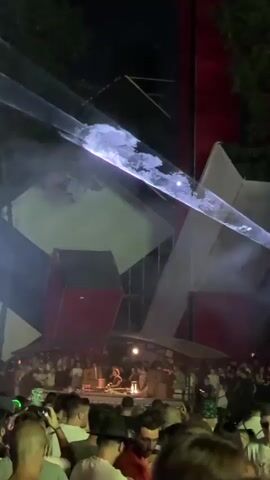 A nighttime outdoor festival scene is depicted with a large crowd gathered in front of a stage. The stage features abstract geometric structures with red and white panels. Laser lights in white and green pierce the smoky atmosphere, creating dramatic beams and patterns. People in the foreground, some shirtless, are looking towards the stage, with some holding phones. The overall atmosphere suggests a lively music event.
