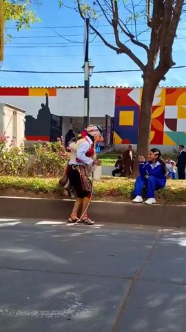 A young girl in a blue tracksuit sits on a curb, holding a smartphone and taking a picture of a person in a traditional costume. The costumed person is leaning forward, facing the girl, and appears to be interacting with her. The costume is elaborate, featuring a mask, a headdress, colorful fabrics, and tassels. The background shows a building with a vibrant mural and a tree. Other people are visible in the background, including a man in a suit. The scene takes place outdoors on a sunny day.