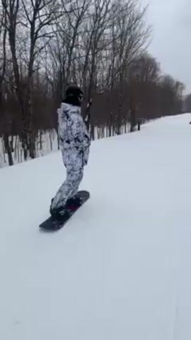 A person in a white and gray camo snowsuit and black helmet is snowboarding down a snowy slope on a cloudy day. The snowboard is black and has the word "Salomon" on it. There are trees on the side of the slope and another person snowboarding further down the slope. The camera angle is from behind and then switches to a first-person view from above the snowboard.