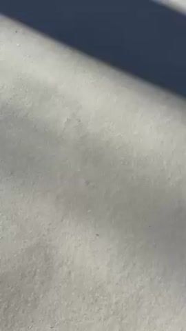 A close-up video shows a hand drawing a heart shape on a snowy surface. The hand has red nail polish and is wearing a black coat. The snow appears to be lightly textured and a shadow covers the upper part of the surface. The hand traces a heart outline in the snow, starting on the right side and moving counter-clockwise to complete the shape.