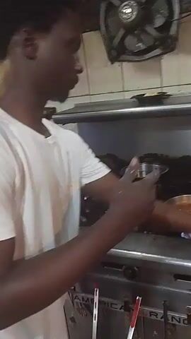 The video shows a person in a white t-shirt, with dark skin and short black hair, standing in front of a stainless steel commercial range. The person is wearing a white t-shirt. The person is holding a pair of metal tongs and is actively cooking something in a pan on the stovetop. There are at least three pans on the stove, with flames visible under one of them. The background appears to be a commercial kitchen, with tiled walls and stainless steel appliances. The person is focused on the cooking process, stirring or manipulating the contents of the pan with the tongs. The lighting is dim, suggesting the video was taken in a kitchen environment.