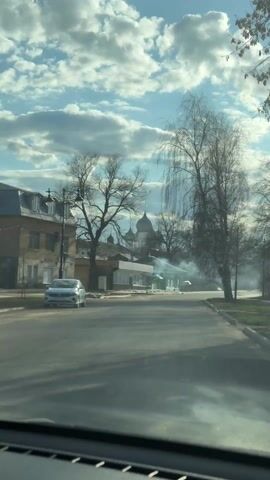 The video, filmed from inside a car, shows a street view on a sunny day with scattered clouds. In the distance, an Orthodox church with dark domes is visible. The street is lined with bare trees and buildings, one of which appears to be a store. Smoke or mist hangs in the air near the church. A car is parked on the left side of the street. The overall scene has a tranquil, slightly hazy atmosphere.