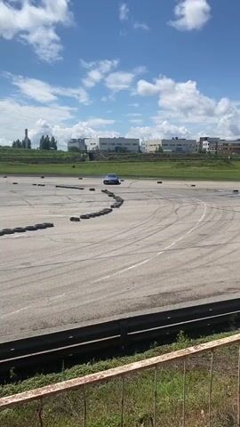 The video shows a blue car drifting on a paved track. The car is in the center of the frame and is kicking up smoke from its tires. There is another white car in the background on the left side of the frame. The track is surrounded by a grassy area and a fence. In the distance, there are buildings and a tall tower. The sky is blue with white clouds. There is a person walking in the background in front of the blue car on the right side of the frame. The car is moving in a counter-clockwise direction.