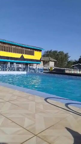 The video shows an outdoor swimming pool on a sunny day. The pool is rectangular and filled with clear blue water, with several people swimming in it. The camera is initially focused on the pool and then shifts to show the feet of a person relaxing on a lounge chair by the pool. A building with yellow and blue accents stands next to the pool, featuring a sign that reads "OGELEA KWA RAHA ZAKO". The surrounding environment includes trees and smaller structures, suggesting a recreational or resort setting. The camera appears to be handheld, with slight movements throughout the video.