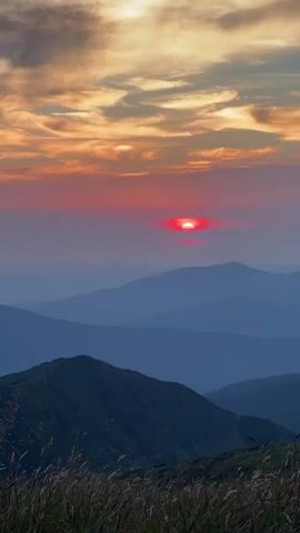 The video shows a time-lapse of a sunset over mountains. The sun is a bright red orb, partially obscured by clouds. The mountains are layered in shades of blue and purple, with some vegetation in the foreground. The sky is filled with orange and pink clouds. There is no audible dialogue or sound. The video appears to be a still image with no movement.