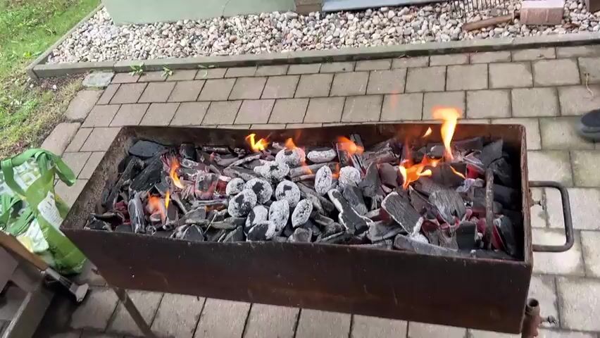 The video shows a long, rectangular barbecue grill filled with burning charcoal. Flames are visible in several areas of the grill, indicating that the charcoal is actively burning. Some of the charcoal pieces are covered in white ash, suggesting they have been burning for some time. The grill is positioned outdoors on a paved patio area. To the left of the grill, a green bag, possibly containing more charcoal, is visible, along with some barbecue tools. In the background, there is a gravel area and a building structure. The lighting suggests it is daytime. There are no people clearly visible in the frame, though a person's foot wearing a dark shoe is partially visible on the right side of the image. The video appears to be a static shot focused on the burning charcoal.