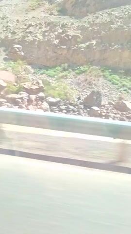 The video shows a view from a moving vehicle on a road running alongside a rocky hillside. The hillside is composed of various sizes of rocks and gravel, with sparse vegetation. A metal guardrail is visible between the road and the hillside. The sky is partly cloudy. The road surface is gray asphalt.