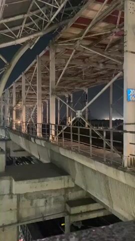 The video depicts an elevated train track structure at night. The structure is made of metal beams and concrete supports. A train, blue and silver in color, is traveling along the track. The lights of a distant cityscape are visible in the background. People are visible inside the train. The camera is positioned below the train track, looking up at the passing train.