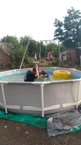 A person in a black shirt is in a round above-ground swimming pool. They are splashing water upwards with their hair. A yellow inflatable ring is floating in the pool to the right of the person. The pool is set up outdoors with trees and structures in the background.