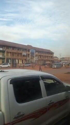 The video begins with a view of a long, multi-story building with a reddish-brown tiled roof. The building appears to have shops or businesses on the ground floor, many of which are closed or covered. There are several cars, motorcycles, and a three-wheeled vehicle (likely an auto-rickshaw or tuk-tuk) on a dirt road in front of the building. Several people are visible, some walking and others standing in small groups. The sky is partly cloudy. The camera is mounted on a white pickup truck, and the edge of the truck's bed and taillight are visible in the foreground. As the video progresses, the camera moves slightly, showing more of the street and the vehicles. The overall impression is of a busy street scene in what appears to be a town or city, possibly in a region with red soil.