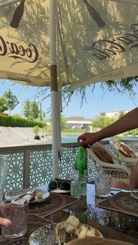 The video captures an outdoor dining scene at a restaurant or cafe, likely beside a river or canal. In the foreground, a dark table is set with patterned placemats, glasses, plates with food (including dumpling-like items), and cutlery. A pair of sunglasses rests on the table. A hand is seen pouring water from a green bottle into a glass.

Above the table, a large, light-colored umbrella with the "Coca-Cola" logo provides shade. Beyond the table, a decorative metal railing with a geometric pattern separates the dining area from a body of water. On the far side of the water, lush green trees and foliage line the banks, with several buildings visible in the distance, including a taller, modern structure and a bridge. Wicker chairs are placed around the table, and a person is partially visible in one of them in the background.

The scene is brightly lit by natural sunlight, indicating midday or early afternoon, with clear skies. The overall atmosphere appears relaxed, suggesting a pleasant meal in an open-air setting. The user context identifies the location as Kuchtumgali, Uzbekistan.