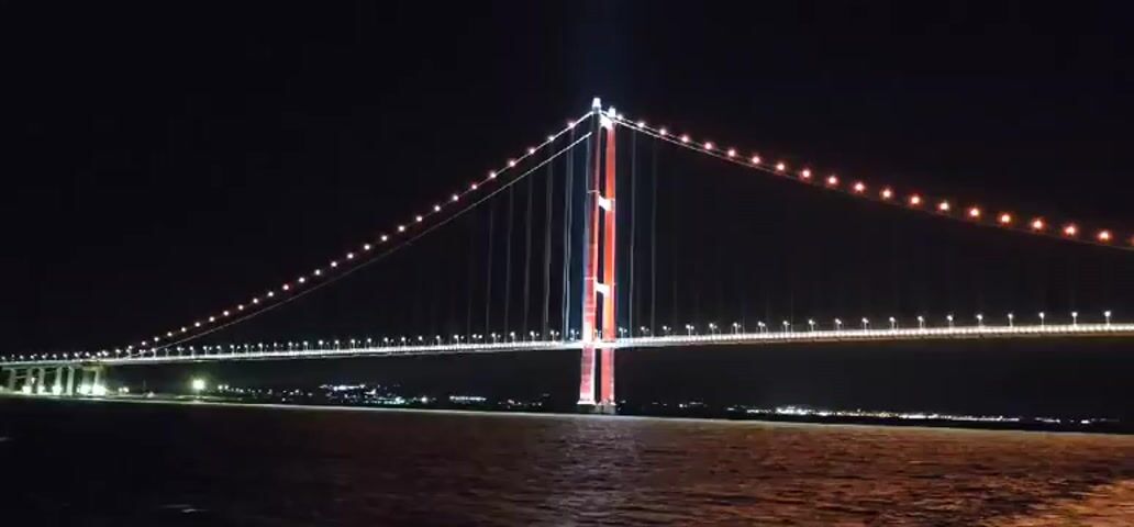 This video captures a suspension bridge at night, illuminated by numerous lights. The bridge spans across a body of water, with city lights visible in the distance along the shoreline. The camera appears to be moving, causing a slight blur in the image, especially on the lights of the bridge and the distant city. The water in the foreground reflects the lights from the bridge and the city, creating shimmering patterns. The sky is dark, indicating nighttime. The bridge is the central focus of the video, with its imposing structure and bright illumination highlighting its architectural features. The lighting on the bridge's towers is particularly striking, with vertical red and blue lights accentuating their height. The suspension cables are lined with small, white lights, creating a sense of scale and detail. The overall scene conveys a sense of nighttime grandeur and technological achievement.