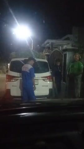 A group of people are standing near a white car at night. A person in a blue uniform is standing on the left of the car. Two other people are standing to the right of the car. One person is wearing a dark hoodie and jeans, and the other is wearing a green shirt and a patterned skirt. The person in the hoodie is leaning against the car, and the person in the green shirt is standing next to a gate. A bright light is visible in the background, illuminating the scene.