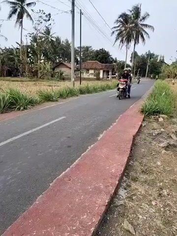 A motorcycle carrying two people drives down a paved road. The person in front wears a black jacket and jeans, and the person behind wears a pink shirt, jeans, and a red backpack. Both wear helmets. The motorcycle is red. To the left of the road, a person on another motorcycle rides away from the camera. This person wears a white headscarf and a dark jacket, and has a black bag on the back of their motorcycle. The road is lined with grass and palm trees. In the background, there are houses and fields. The sky is hazy.