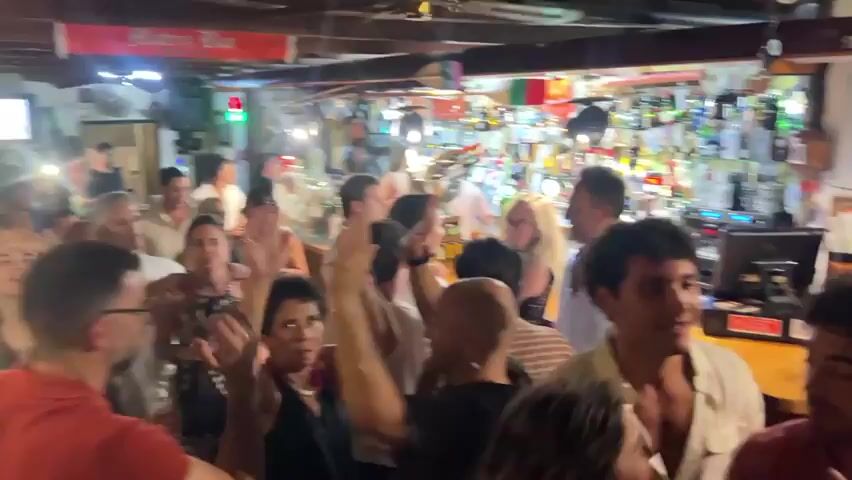 The video shows a group of people in what appears to be a dimly lit indoor venue, possibly a restaurant or bar. The camera is shaky and the footage is blurry, making it difficult to identify specific individuals or details clearly. In the foreground, several people are visible, their faces mostly obscured or out of focus. One person, in the center of the frame, is wearing a pink shirt. To the left, a person in a light-colored shirt is looking towards the right. In the background, a bar area is visible with shelves stocked with bottles. There is also a framed picture on the wall and some neon lighting. The overall atmosphere seems to be social and lively, with people engaged in conversation or movement.