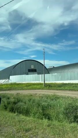 A wide shot captures a long, arched, metal building under a partly cloudy blue sky. The building is constructed of corrugated metal panels and has a white base. The surrounding area is dry and grassy, with some yellow flowers visible. A dirt road runs in front of the building, and a faint line of what appears to be train tracks is also visible. A utility pole stands in the foreground on the right side of the frame. The video appears to be shot from a moving vehicle, indicated by the slight motion blur and the reflection of a car mirror on the left side of the frame.