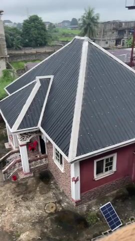 The video pans across a residential area, showcasing a red house with a dark metal roof. The yard is unpaved, with some scattered debris and a small solar panel. In the background, an unfinished multi-story building is visible, along with lush greenery and other structures. Two motorcycles are parked in the yard. The sky appears overcast.